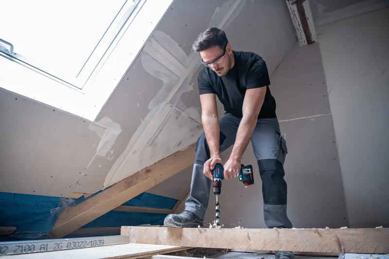 บุคคลที่สวมอุปกรณ์ด้านความปลอดภัยกำลังเจาะเข้าไปในคานไม้ในห้องใต้หลังคาที่ตกแต่งเสร็จเพียงบางส่วน.