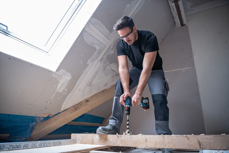 บุคคลที่สวมอุปกรณ์ด้านความปลอดภัยกำลังเจาะเข้าไปในคานไม้ในห้องใต้หลังคาที่ตกแต่งเสร็จเพียงบางส่วน.