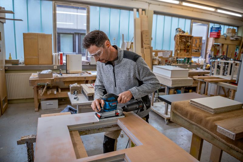 บุคคลที่สวมอุปกรณ์ป้องกันความปลอดภัยกำลังขัดไม้ด้วยเครื่องขัดกระดาษทรายแบบสั่นในโรงงาน