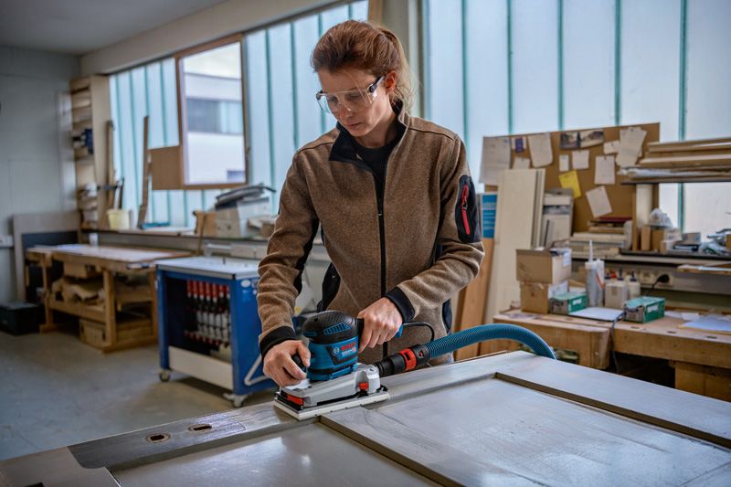 บุคคลที่สวมอุปกรณ์ด้านความปลอดภัยกำลังขัดประตูไม้ด้วยเครื่องขัดกระดาษทรายแบบสั่นในโรงงาน