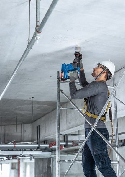 บุคคลที่สวมอุปกรณ์ด้านความปลอดภัยเจาะเพดานคอนกรีตขณะยืนอยู่บนนั่งร้าน.