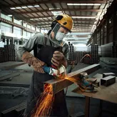 บุคคลที่สวมอุปกรณ์ป้องกันความปลอดภัยกำลังใช้เครื่องเจียรเจียรบดคานโลหะ จนเกิดประกายไฟพุ่งออกมา.