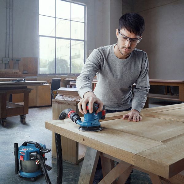 บุคคลที่สวมอุปกรณ์ด้านความปลอดภัยกำลังขัดประตูไม้ด้วยเครื่องขัดกระดาษทรายแบบวงกลมที่เชื่อมต่ออยู่.