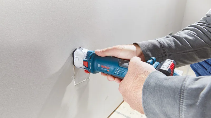 คนๆ หนึ่งกำลังเจาะรูสี่เหลี่ยมบนแผ่น drywall โดยใช้เครื่องเราเตอร์ drywall แบบไร้สาย.