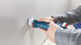 คนๆ หนึ่งกำลังเจาะรูสี่เหลี่ยมบนแผ่น drywall โดยใช้เครื่องเราเตอร์ drywall แบบไร้สาย.