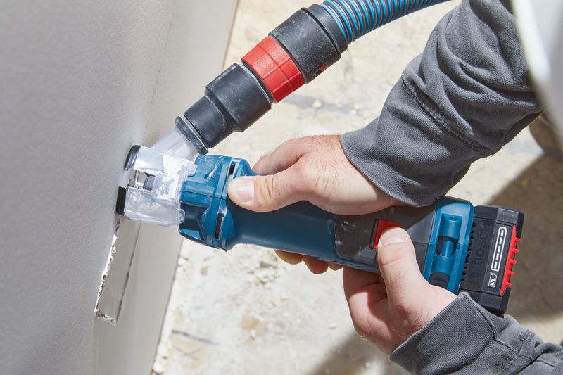 คนๆ หนึ่งกำลังเจาะรูสี่เหลี่ยมบนแผ่นผนัง drywall โดยใช้เครื่องมือไฟฟ้าพร้อมเครื่องดูดฝุ่น.
