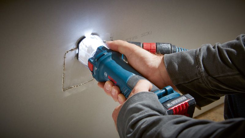 คนๆ หนึ่งกำลังเจาะรูสี่เหลี่ยมบนแผ่น drywall โดยใช้เครื่องเราเตอร์ drywall แบบไร้สาย.