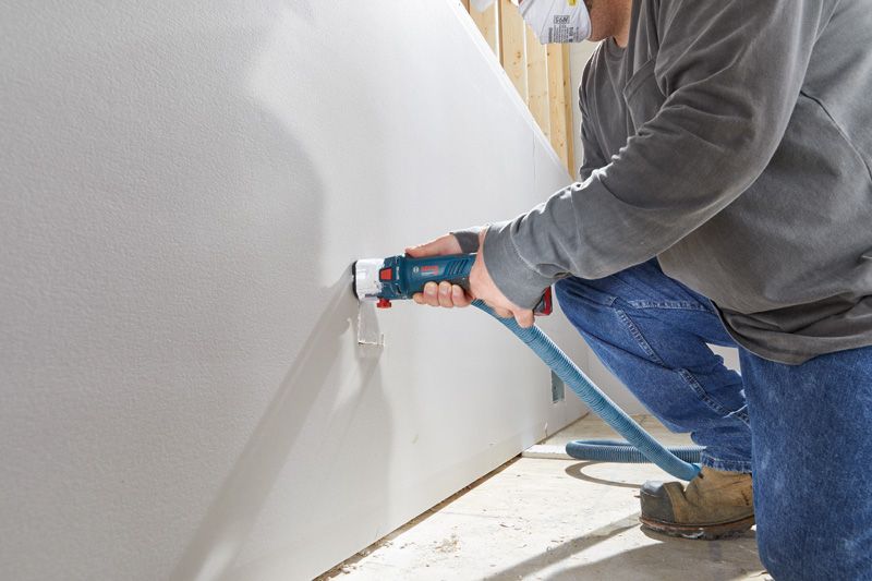 บุคคลที่สวมอุปกรณ์ด้านความปลอดภัยใช้เครื่องเราเตอร์ drywall แบบไร้สายในการตัดช่องเปิด.