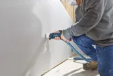 บุคคลที่สวมอุปกรณ์ด้านความปลอดภัยใช้เครื่องเราเตอร์ drywall แบบไร้สายในการตัดช่องเปิด.