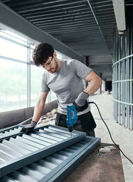 บุคคลที่สวมอุปกรณ์ความปลอดภัยกำลังเจาะสกรูเข้ากับโครงโลหะบนไซต์ก่อสร้าง.