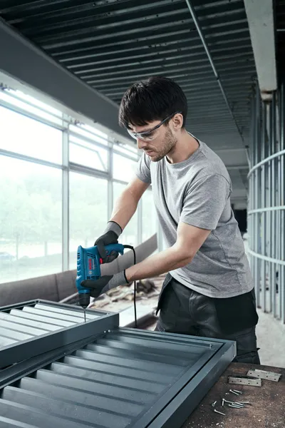 บุคคลที่สวมอุปกรณ์ด้านความปลอดภัยใช้สว่านไฟฟ้าในการประกอบโครงโลหะ.