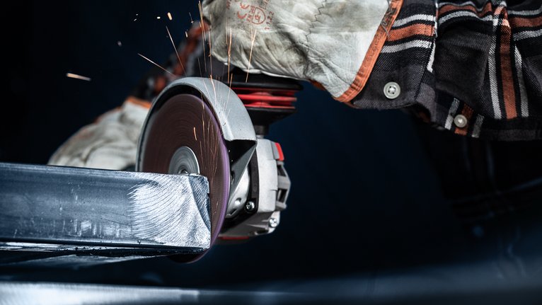 บุคคลที่สวมอุปกรณ์ความปลอดภัยกำลังบดคานโลหะด้วยเครื่องมือไฟฟ้า