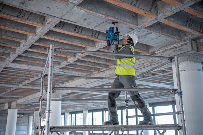 บุคคลที่สวมอุปกรณ์ความปลอดภัยกำลังเจาะเข้าไปในเพดานคอนกรีตในพื้นที่ก่อสร้าง.