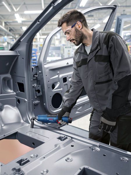 บุคคลที่สวมอุปกรณ์ด้านความปลอดภัยใช้เครื่องมือไฟฟ้าบนโครงรถยนต์ในสถานประกอบการอุตสาหกรรม.