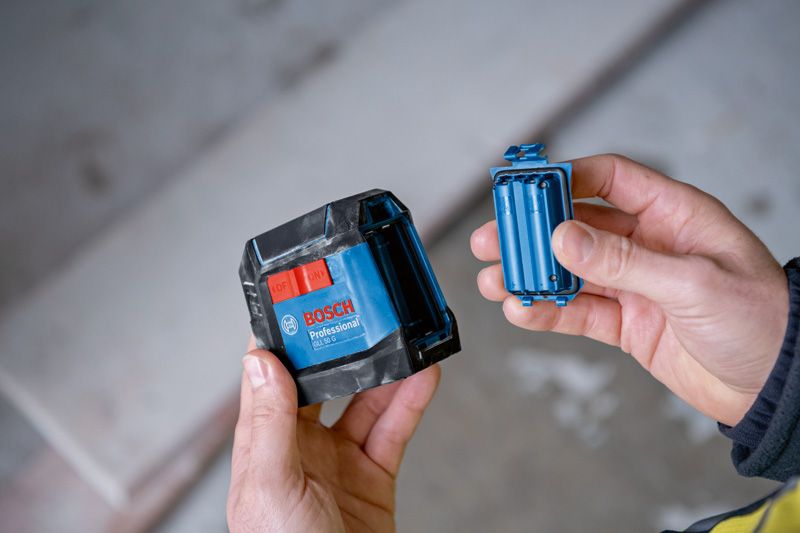 บุคคลถือเครื่องมือวัดระดับเลเซอร์สีน้ำเงินและช่องใส่แบตเตอรี่ในบ้าน.