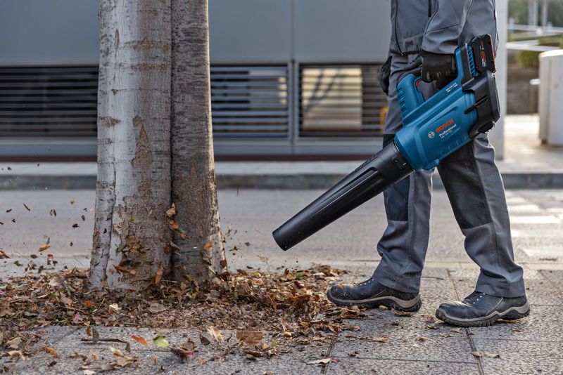 บุคคลที่สวมอุปกรณ์ด้านความปลอดภัยใช้เครื่องเป่าใบไม้ไร้สายเพื่อเคลื่อนย้ายใบไม้ใกล้ต้นไม้