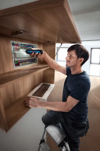 คนบนบันไดใช้ไขควงไร้สายทำงานกับแผงควบคุมไฟฟ้า.