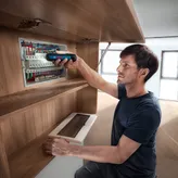 คนๆ หนึ่งกำลังยึดสายไฟในแผงไฟฟ้าโดยใช้ไขควงไร้สาย.