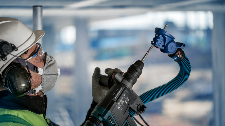 บุคคลที่สวมอุปกรณ์ด้านความปลอดภัยเจาะเข้าไปในเพดานโดยใช้เครื่องมือไฟฟ้าพร้อมเครื่องดูดฝุ่น