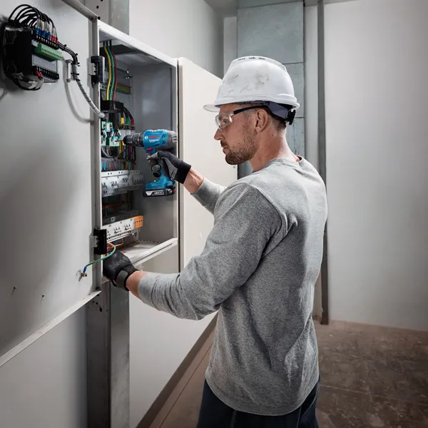 บุคคลที่สวมอุปกรณ์ด้านความปลอดภัยใช้สว่านไร้สายบนแผงควบคุมไฟฟ้า.