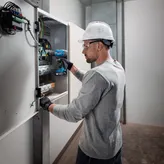 บุคคลที่สวมอุปกรณ์ด้านความปลอดภัยใช้สว่านไร้สายบนแผงควบคุมไฟฟ้า.