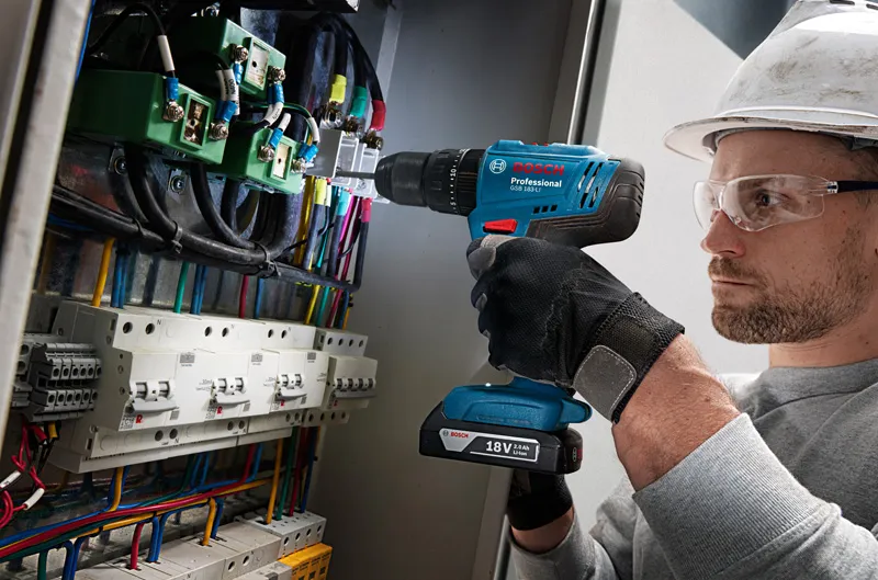 บุคคลที่สวมอุปกรณ์ความปลอดภัยกำลังเจาะเข้าไปในแผงควบคุมที่เต็มไปด้วยสายไฟ.