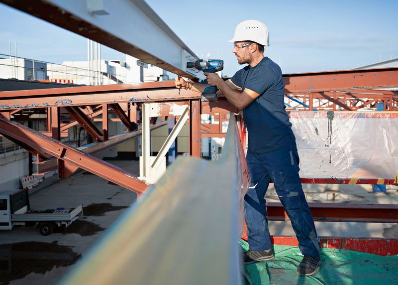 บุคคลที่สวมอุปกรณ์ด้านความปลอดภัยใช้ประแจกระแทกไร้สายกับคานเหล็ก.