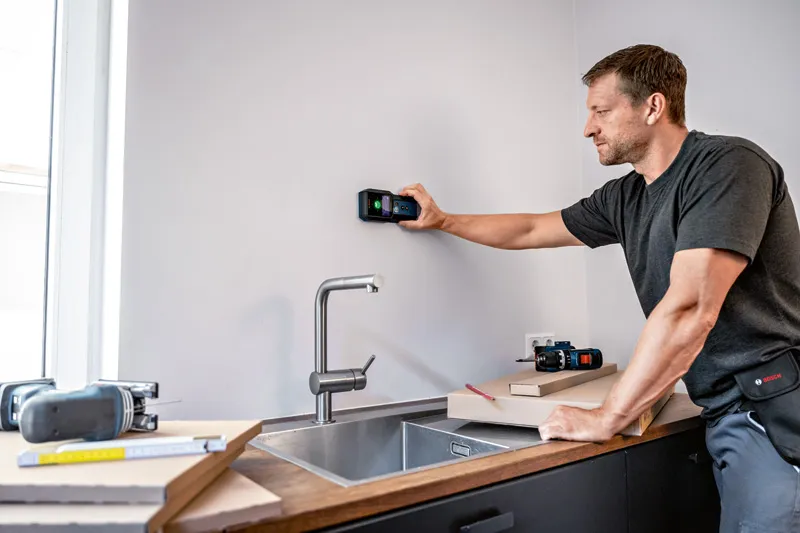 คนๆ หนึ่งกำลังสแกนผนังห้องครัวเหนืออ่างล้างจานโดยใช้เครื่องตรวจจับแบบดิจิทัล.