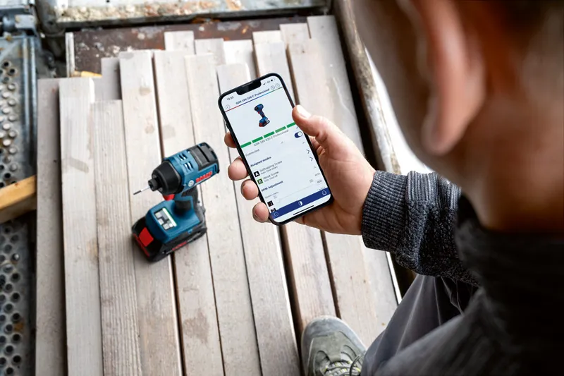 บุคคลกำลังตรวจสอบฟีเจอร์แอปของเครื่องมือไฟฟ้าบนสมาร์ทโฟนใกล้กับสว่านไร้สาย.