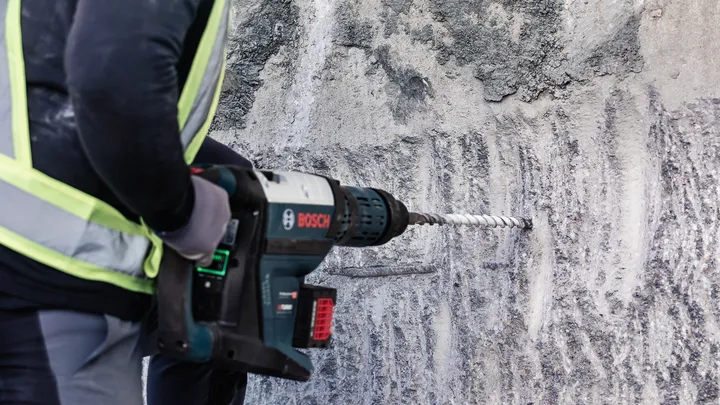 บุคคลที่สวมอุปกรณ์ความปลอดภัยกำลังเจาะเข้าไปในผนังคอนกรีตโดยใช้เครื่องมือไฟฟ้า