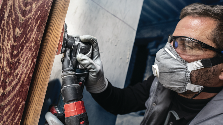 บุคคลที่สวมอุปกรณ์ด้านความปลอดภัยกำลังขัดพื้นผิวไม้ด้วยเครื่องมือไฟฟ้า