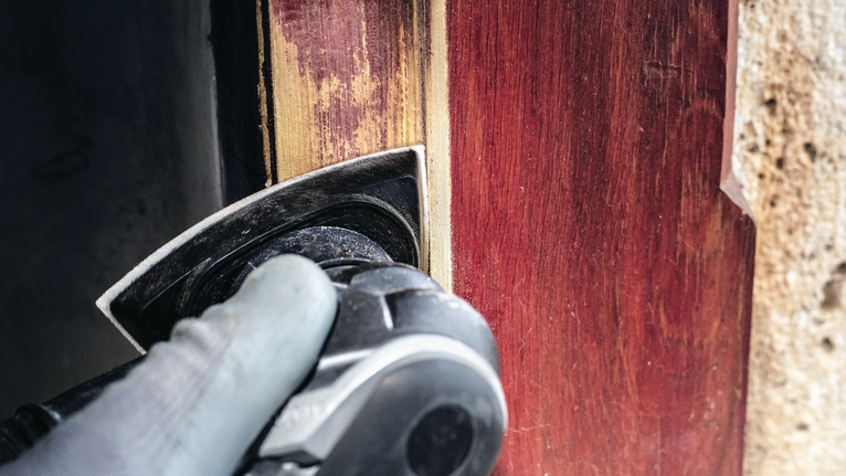 บุคคลที่สวมอุปกรณ์ความปลอดภัยกำลังขัดกรอบประตูไม้ด้วยเครื่องมือไฟฟ้า