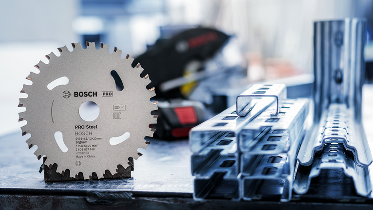 ใบเลื่อยวงเดือนตั้งตรงบนโต๊ะทำงานข้างคานโลหะที่วางซ้อนกัน