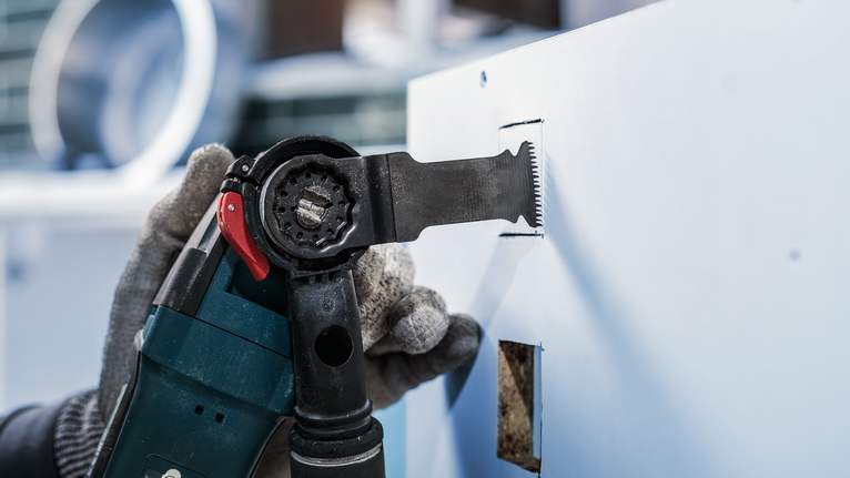 บุคคลที่สวมอุปกรณ์ด้านความปลอดภัยกำลังใช้เครื่องมือไฟฟ้าตัดรูสี่เหลี่ยมบนแผ่นผนังยิปซัม