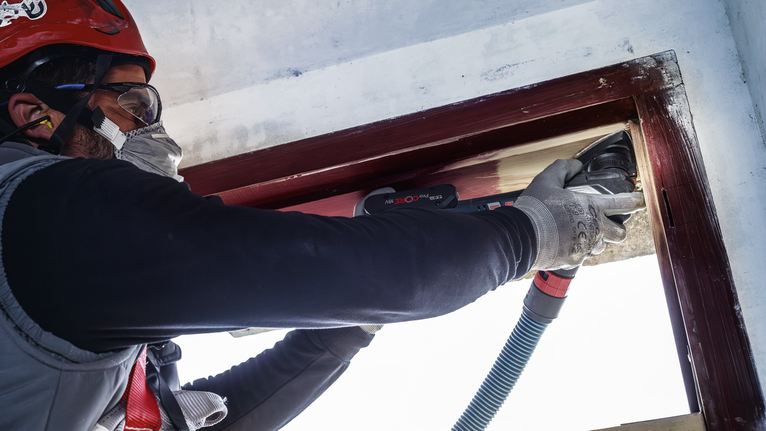 บุคคลที่สวมอุปกรณ์ความปลอดภัยกำลังขัดกรอบหน้าต่างไม้ด้วยเครื่องมือไฟฟ้า