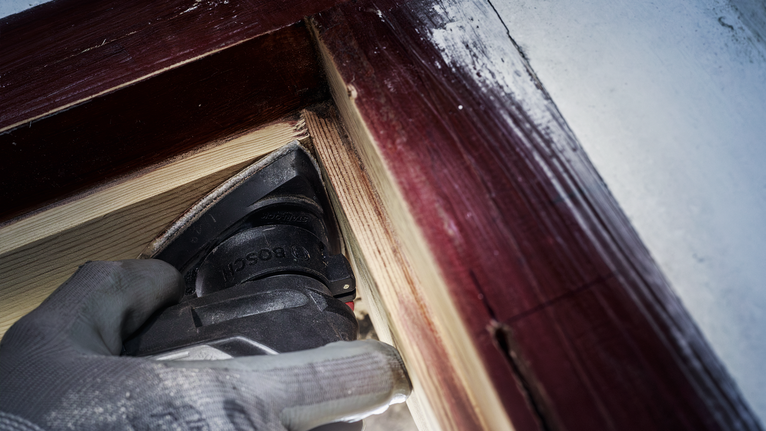 บุคคลที่สวมอุปกรณ์ความปลอดภัยกำลังขัดไม้บริเวณกรอบหน้าต่างที่ทาสี