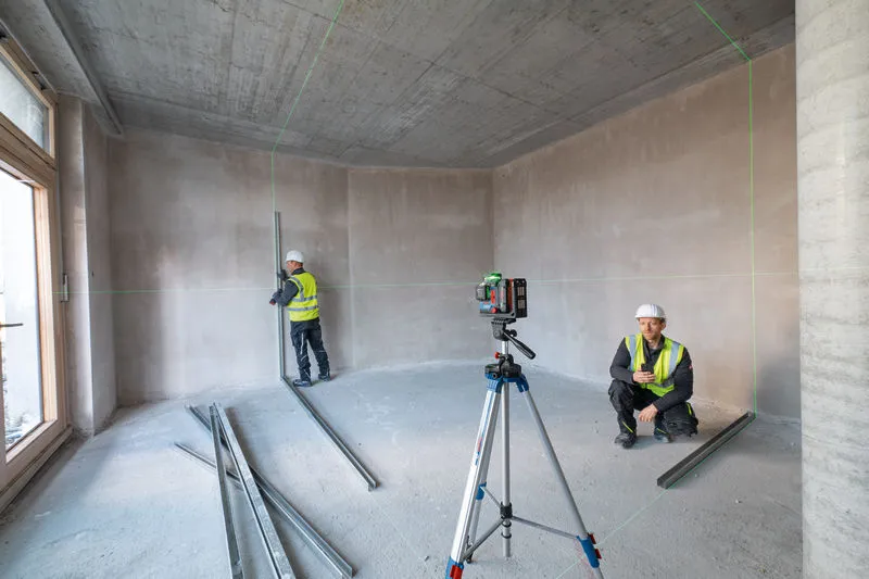 คนสองคนที่สวมอุปกรณ์ความปลอดภัยกำลังใช้เครื่องมือปรับระดับเลเซอร์ในห้องที่ยังไม่เสร็จ