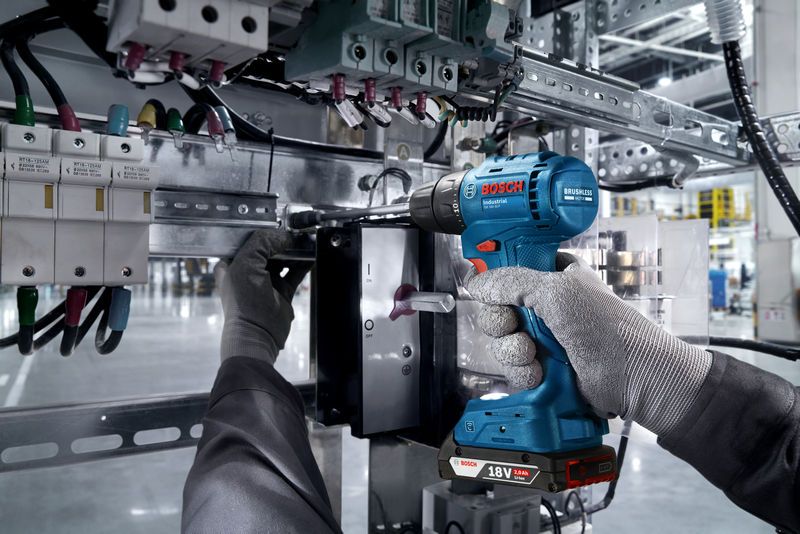 บุคคลที่สวมอุปกรณ์ด้านความปลอดภัยใช้สว่านไร้สายกับแผงควบคุมไฟฟ้า.