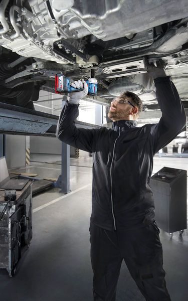 บุคคลที่สวมอุปกรณ์ด้านความปลอดภัยใช้สว่านไฟฟ้าทำงานใต้ตัวรถที่ยกสูง.