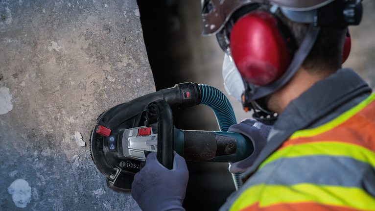คนงานที่สวมอุปกรณ์ความปลอดภัยกำลังขัดคอนกรีตด้วยเครื่องเจียรไฟฟ้ามือถือ.