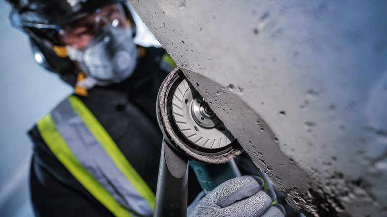 บุคคลที่สวมอุปกรณ์ด้านความปลอดภัยกำลังบดคอนกรีตโดยใช้เครื่องมือไฟฟ้ามือถือ.