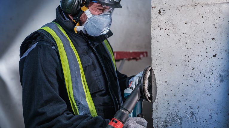 คนงานที่สวมอุปกรณ์ความปลอดภัยกำลังบดผนังคอนกรีตด้วยเครื่องมือไฟฟ้า.