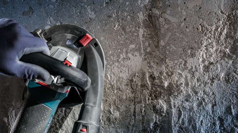 บุคคลที่สวมอุปกรณ์ความปลอดภัยกำลังใช้งานเครื่องมือไฟฟ้าบนผนังคอนกรีตที่ขรุขระ