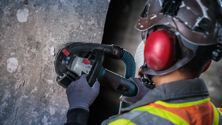 บุคคลที่สวมอุปกรณ์ความปลอดภัยกำลังบดผนังคอนกรีตด้วยเครื่องมือไฟฟ้า