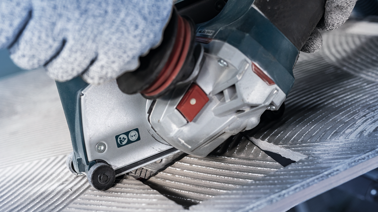 บุคคลที่สวมอุปกรณ์ด้านความปลอดภัยกำลังใช้งานเครื่องมือไฟฟ้าเพื่อตัดพื้นผิวโลหะที่มีร่อง