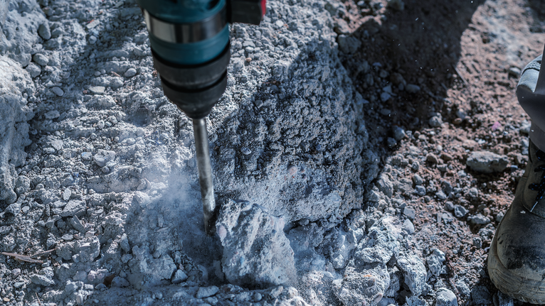 สว่านกระแทกเจาะทะลุคอนกรีตที่ขรุขระ คนงานสวมอุปกรณ์ป้องกันความปลอดภัย.