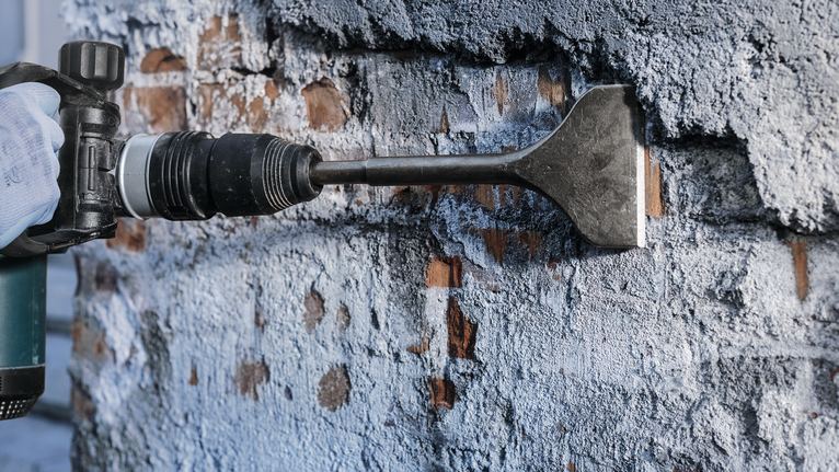 บุคคลที่สวมอุปกรณ์ด้านความปลอดภัยใช้หัวสิ่วเพื่อขูดปูนปลาสเตอร์ออกจากผนัง