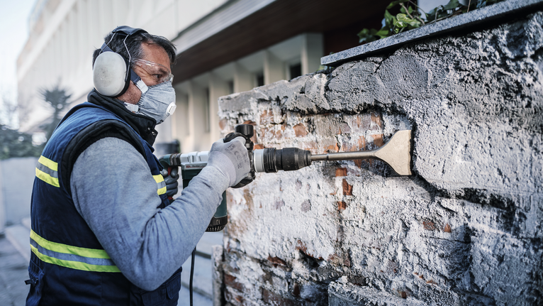 บุคคลที่สวมอุปกรณ์ด้านความปลอดภัยใช้สิ่วไฟฟ้าเพื่อขูดปูนออกจากผนังอิฐ