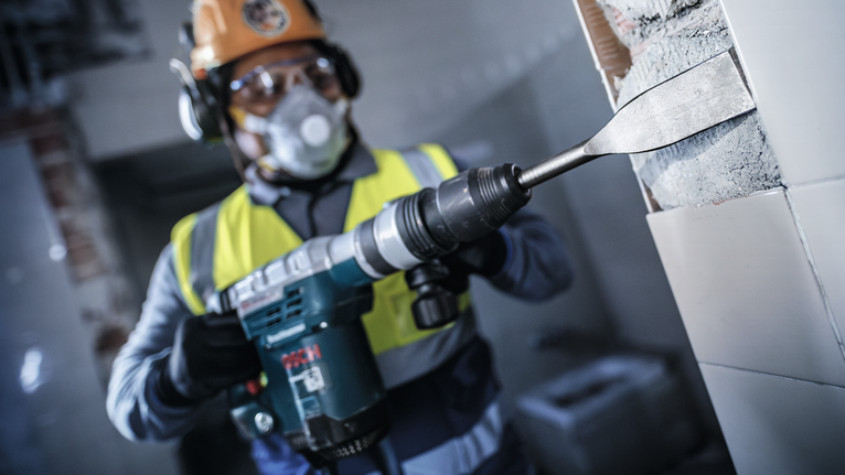 บุคคลที่สวมอุปกรณ์ความปลอดภัยใช้ค้อนโรตารี่ในการสกัดกระเบื้องออกจากผนัง