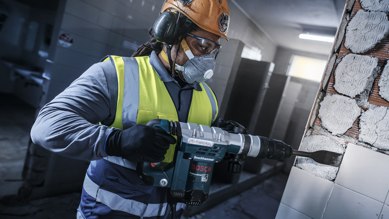 บุคคลที่สวมอุปกรณ์ด้านความปลอดภัยใช้ค้อนโรตารี่เพื่อดึงกระเบื้องออกจากผนัง